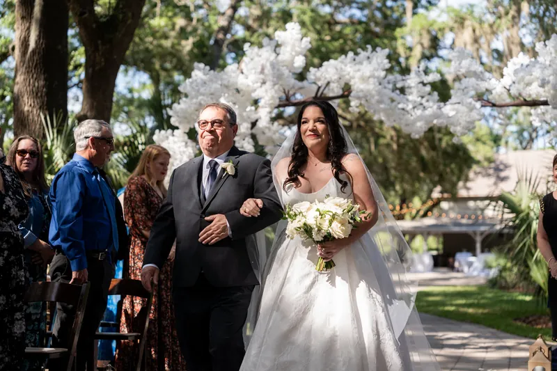 Giselle walks arm-in-arm with her father down the aisle holding white bouquet under blooming white flowers.