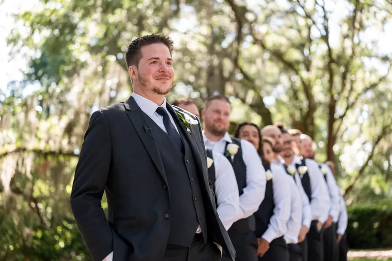 Duncan stands in dark suit with groomsmen lined up behind him before ceremony begins.