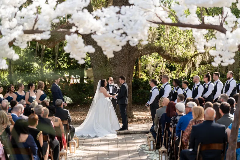 Giselle and Duncan exchange vows under white flowering tree with guests seated on either side.