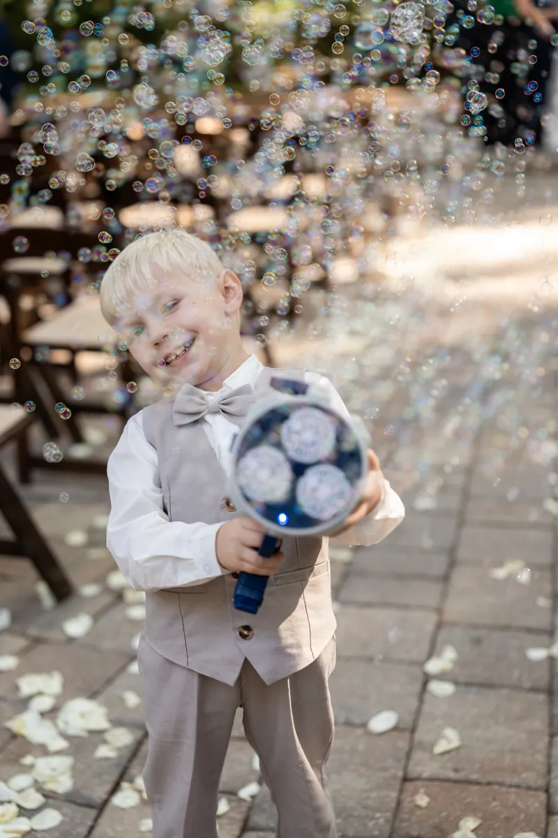 Young ring bearer in a vest holds a blue ring pillow surrounded by falling flower petals.