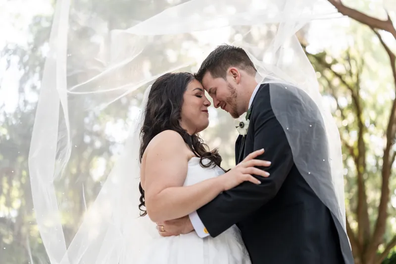Giselle and Duncan embrace face-to-face under flowing veil among tall trees during their wedding ceremony.