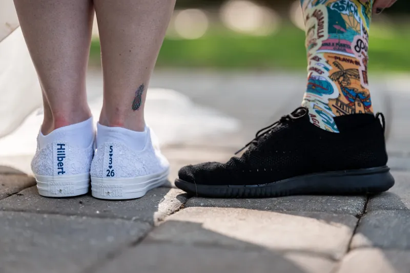 Bride and groom's feet showing Giselle's white sneakers labeled "Bride" and Duncan's black sneakers with colorful socks on pavement.