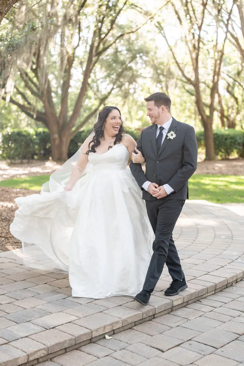 Giselle and Duncan walk together on brick pathway, Giselle's white dress flowing, Duncan in dark suit with hand in pocket.