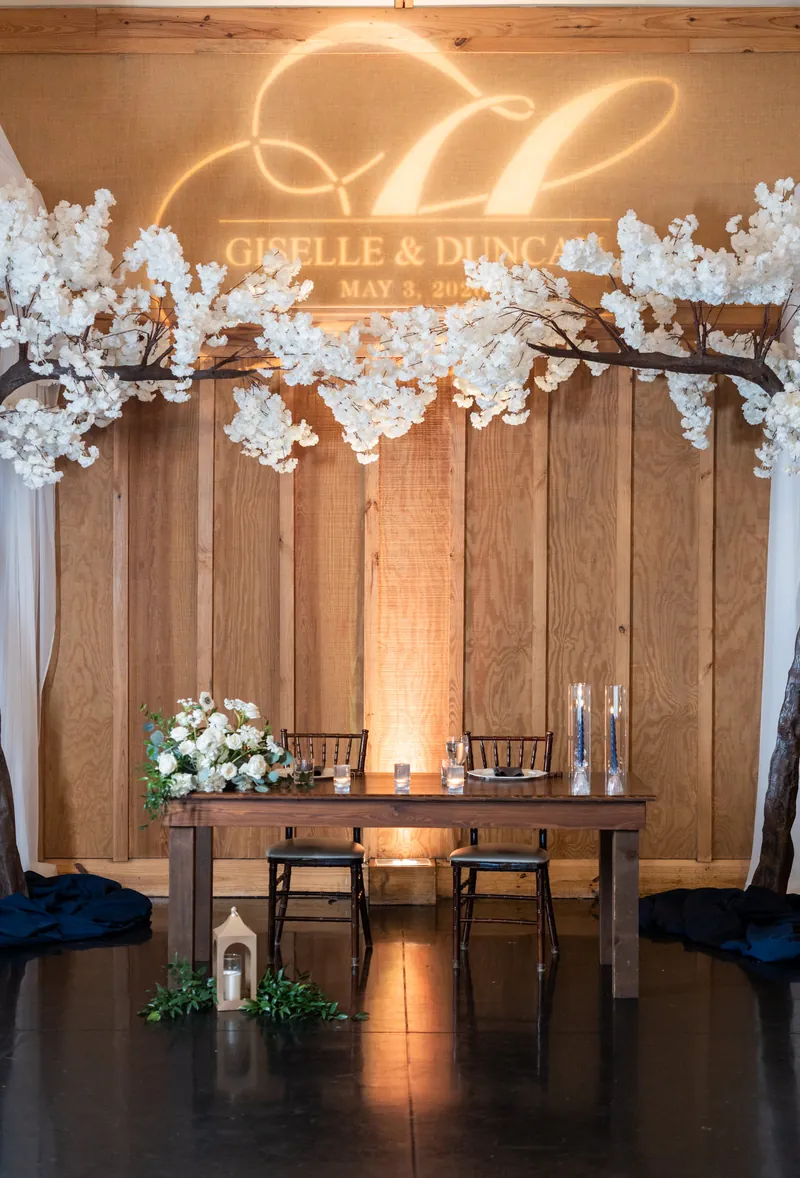 Wooden-backed reception space with neon "G&D" sign above sweetheart table, white floral garland overhead, candlelit ambiance.