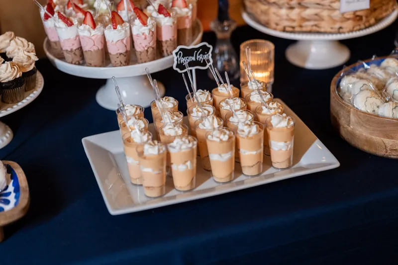 White square platter of caramel-filled dessert cups with striped straws on navy tablecloth surrounded by pastries.