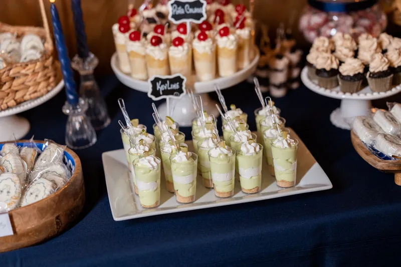 White square platter of green dessert cups and tiered cake with red berries on navy table with multiple desserts.