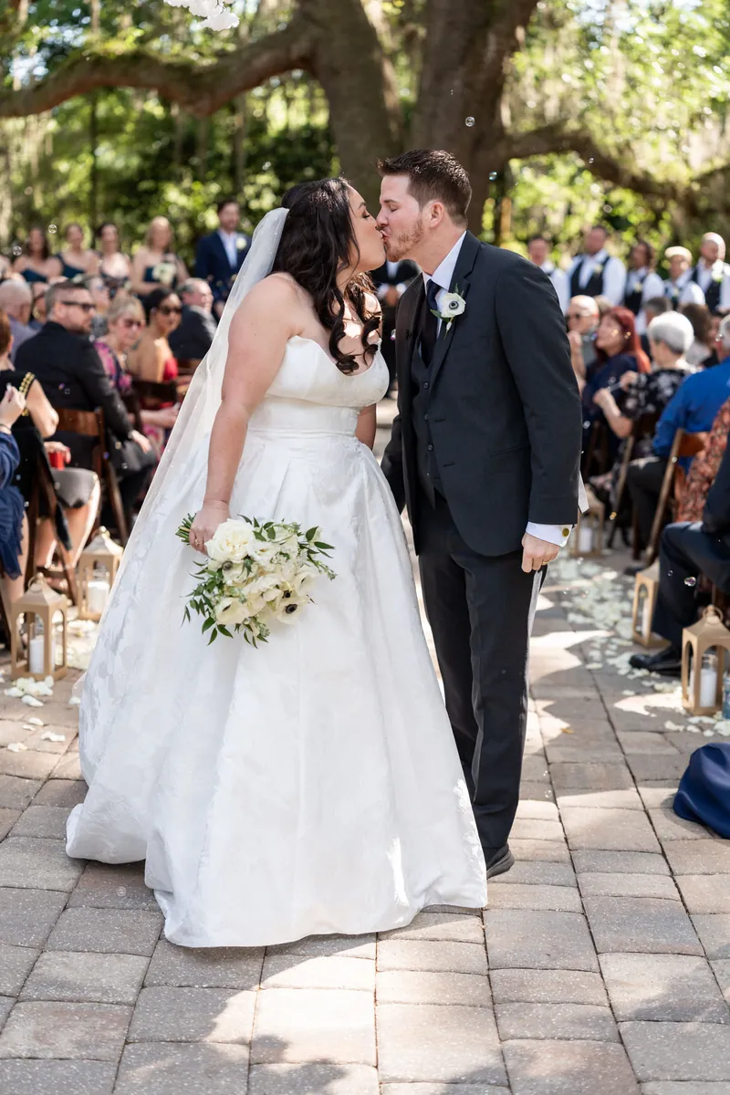 Giselle and Duncan walk down the brick aisle at Bowing Oaks, Giselle holding a white anemone bouquet while guests line