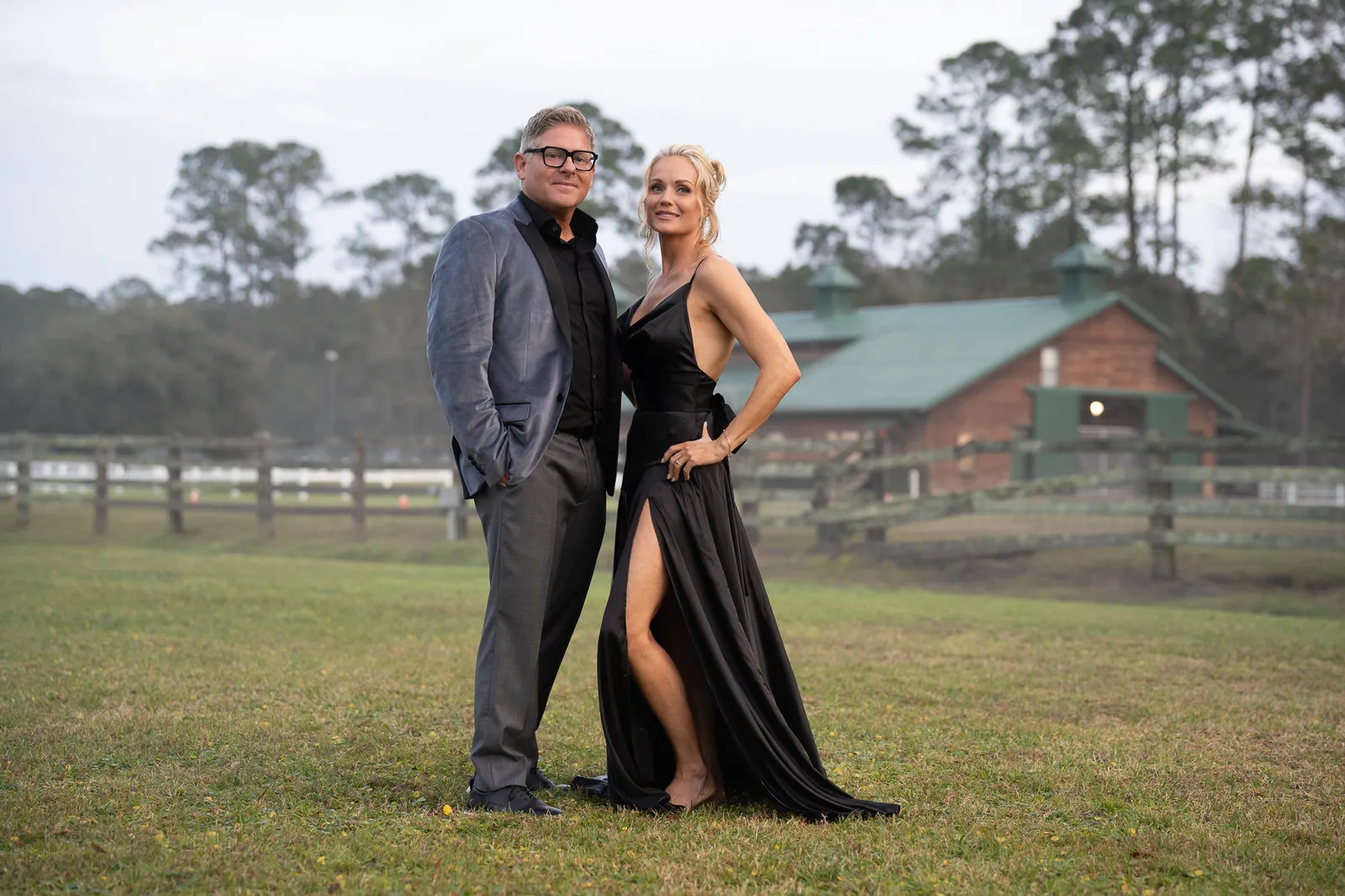 Couple posing elegantly on rural farm property with barn and white fencing