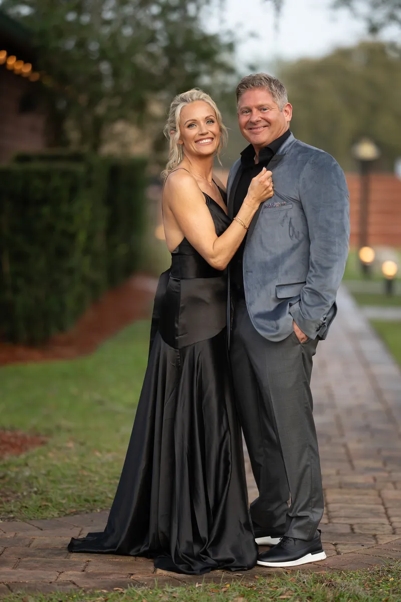 Hamilton wraps his arms around Michelle from behind as they stand outdoors with lit trees visible in the distance.