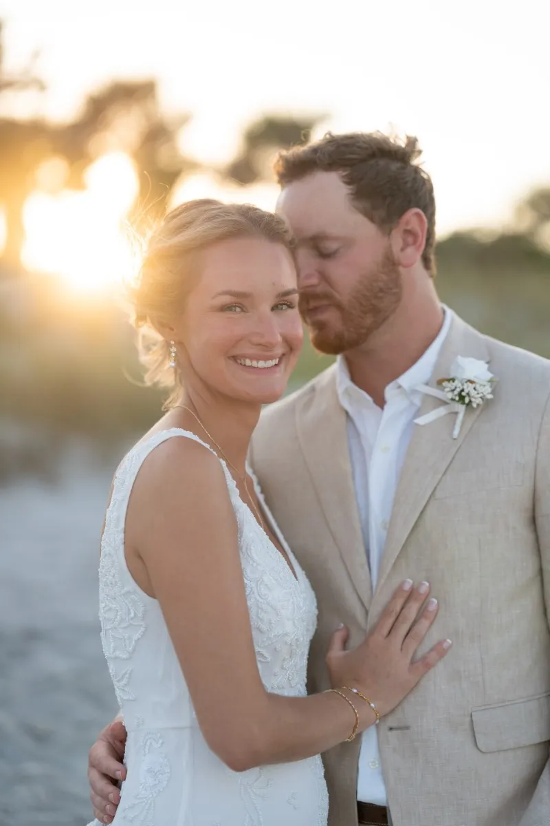 Tanner kisses Harlee's cheek as golden sunlight bathes the beach behind them, both smiling in their wedding attire.
