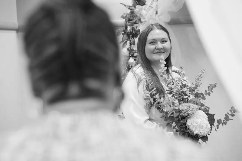 A beautiful black and white portrait captures the bride's joyful smile as she holds her eucalyptus bouquet, with the groom softly blurred in the foreground.