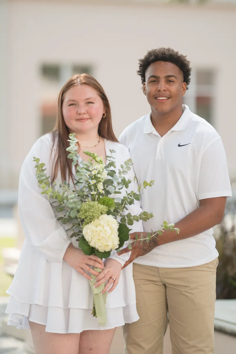 A joyful couple poses together after their courthouse elopement, the bride holding a beautiful white and green bouquet while both smile radiantly in their white attire.