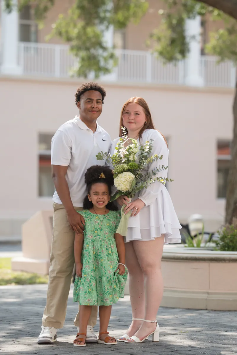 A joyful newlywed couple poses with a young girl after their courthouse elopement ceremony, the bride holding a beautiful bouquet in front of a civic building.