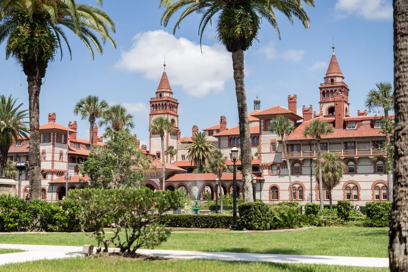 Red-roofed historic mansion with towers framed by palm trees and manicured lawns under blue sky.