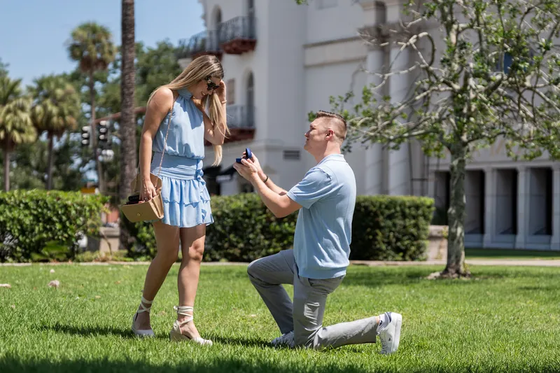 Jakub holds Josy's hand and kneels on one knee as she reacts with joy on the grassy grounds.