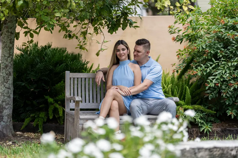 Josy and Jakub sit together on a white bench in a lush courtyard garden surrounded by flowering plants.