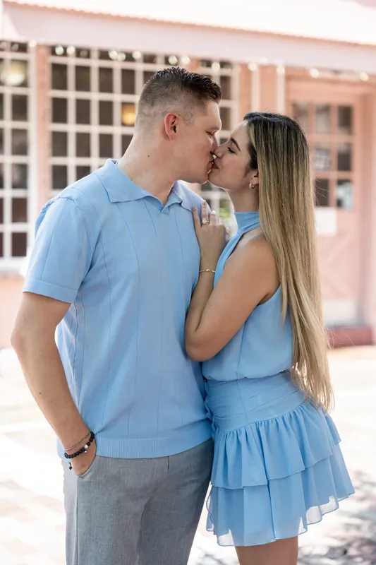 Jakub leans in to kiss Josy's cheek in a sun-dappled courtyard, both dressed in coordinating shades of blue.