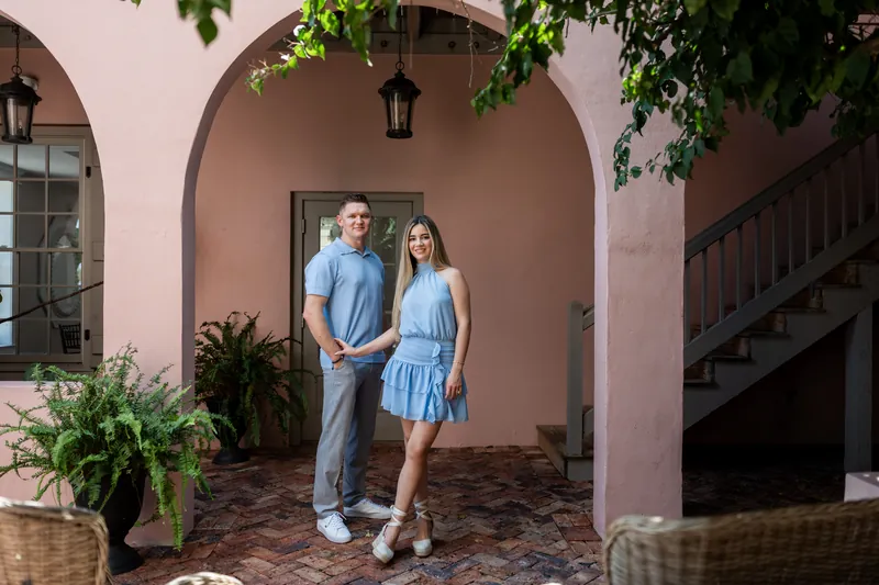 Jakub and Josy stand under a pink arched doorway with ivy, both wearing light blue in a historic courtyard.
