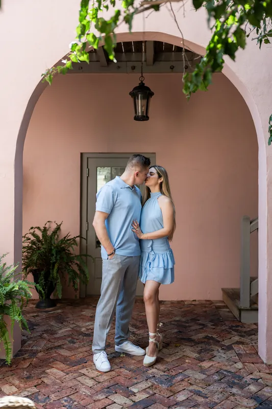 Jakub kisses Josy's forehead as they embrace beneath a terracotta arch with potted plants nearby.