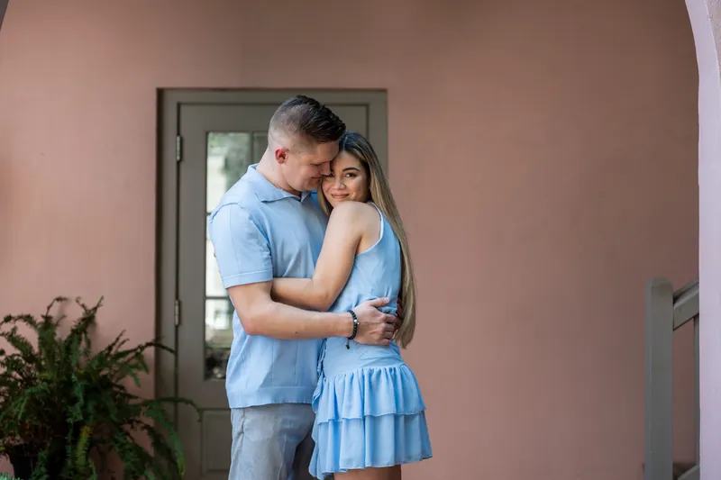Jakub wraps his arms around Josy from behind against a pink stucco wall, both in coordinating blue.