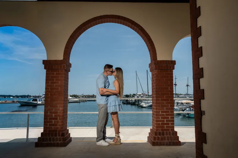 Jakub and Josy embrace under brick archways with sailboats and water visible through the columns behind them.