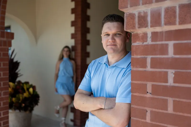 Jakub stands with crossed arms in a light blue shirt against exposed brick, Josy blurred behind him.
