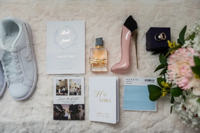 Wedding day details arranged on marble: white sneakers, cologne, pink heels, stationery, and fresh flowers.