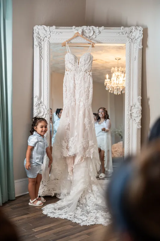 Two young children admire Janet's delicate ivory lace wedding dress hanging in an ornate white-framed mirror.