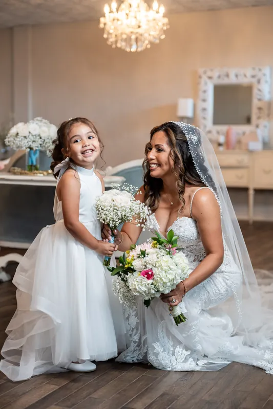 Janet kneels beside a flower girl in white, both smiling together indoors under a chandelier.