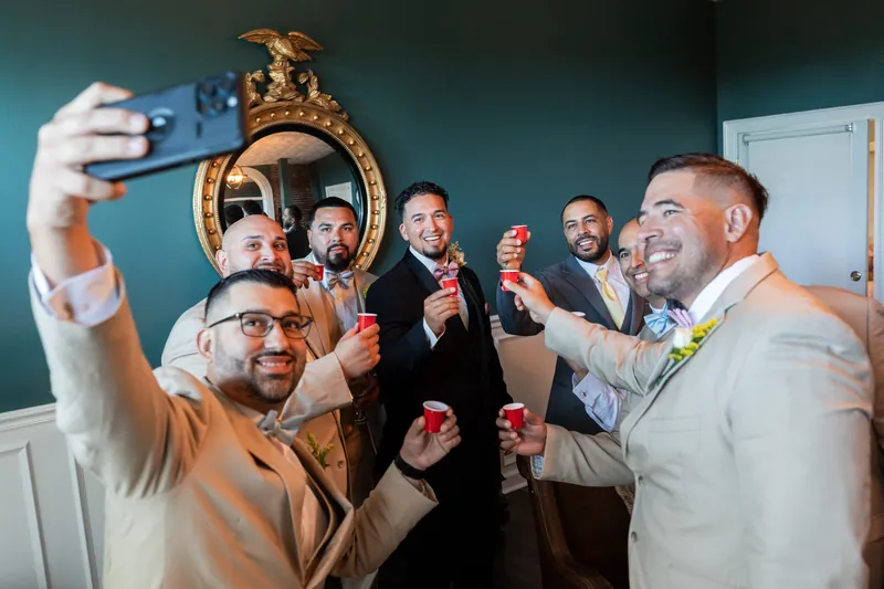 Rick holds up his phone for a selfie with groomsmen gathered around a decorative gold mirror.
