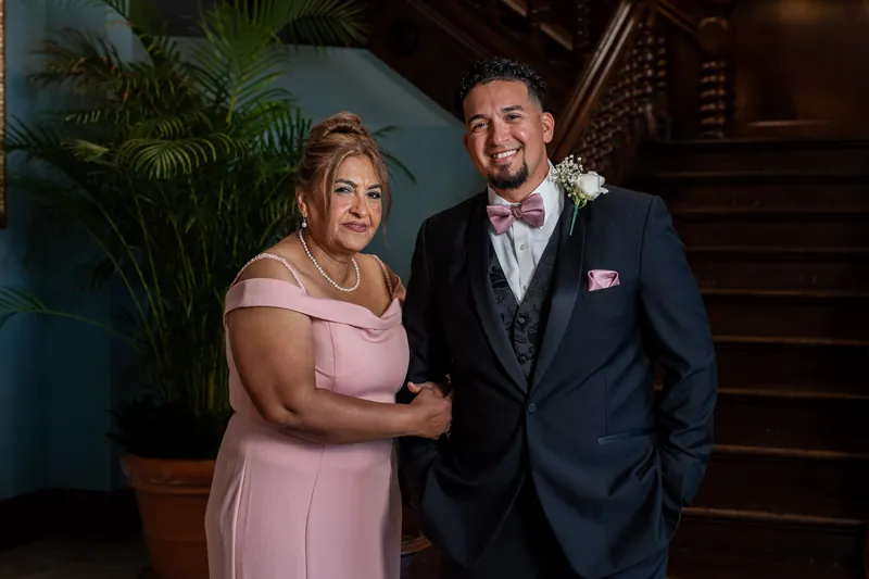 Rick poses with his mother in a pink dress, both smiling under warm lighting near a staircase.