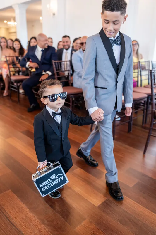 A young boy in a light blue suit walks across the wooden dance floor holding a "here comes the bride" sign.