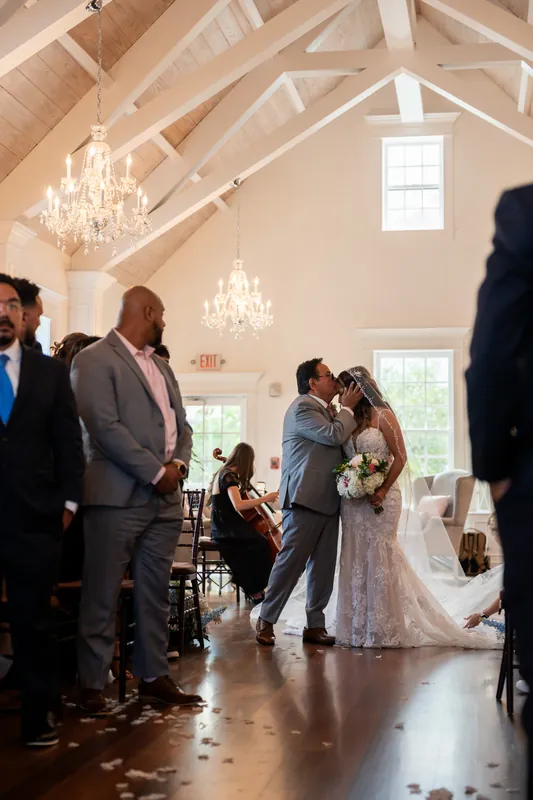 Janet walks toward Rick down the aisle as guests stand and officiant waits beneath ornate chandeliers.