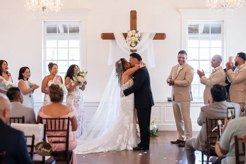 Janet and Rick kiss at the altar as seated guests applaud, with the wooden cross and white columns behind them.