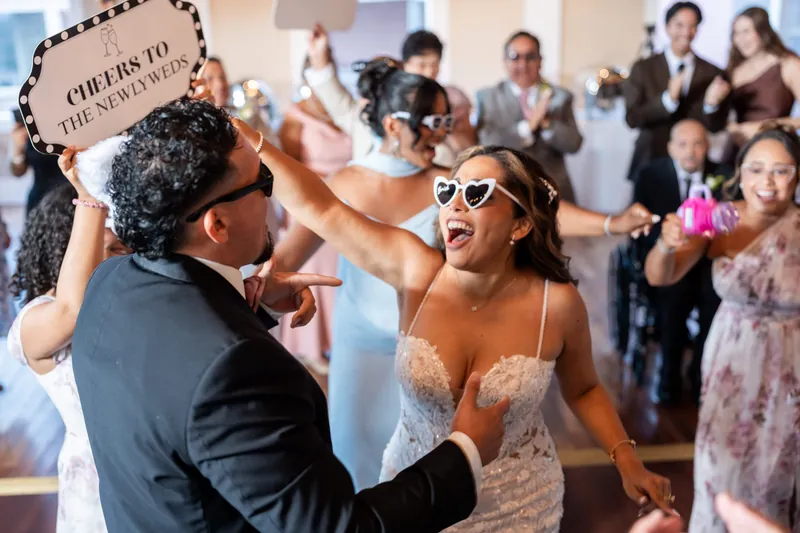 Janet and Rick dance with sunglasses and a prop sign while guests surround them on The White Room's dance floor.