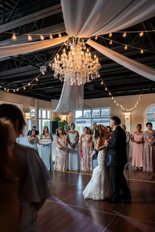 A crystal chandelier hangs beneath draped fabric and string lights as Rick and Janet share their first dance.