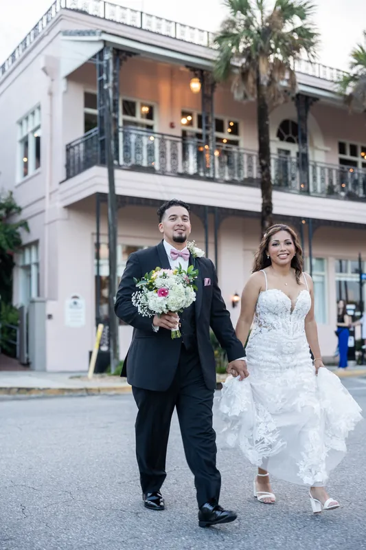 Rick holds Janet's hand and bouquet as they walk down a St. Augustine street in front of a white colonial building.