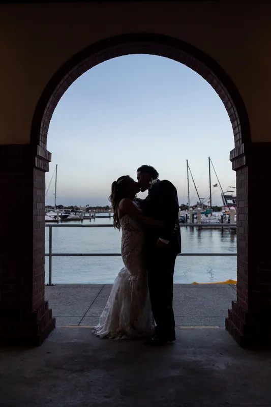 Rick and Janet kiss beneath an arched stone doorway framing sailboats and waterfront beyond.