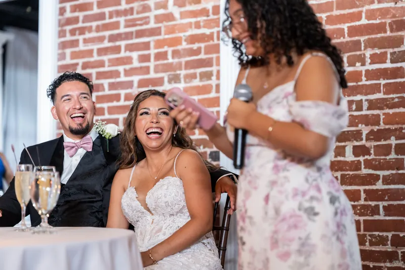 Rick laughs alongside Janet at their reception table as a bridesmaid speaks, with exposed brick visible behind them.