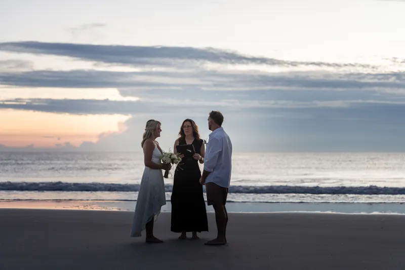 Jarah and Brandon stand with their officiant on wet sand as the sky shifts to dusk.