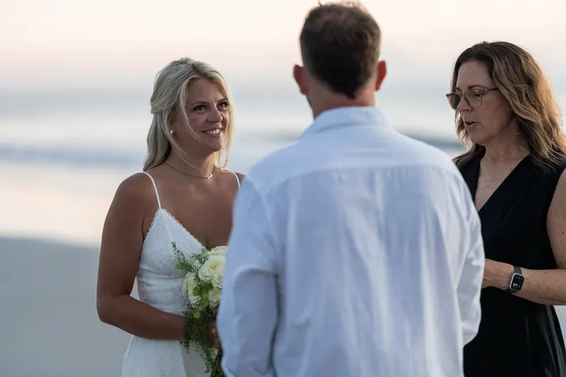 Jarah holds a white bouquet and smiles at Brandon during their ceremony by the shore.