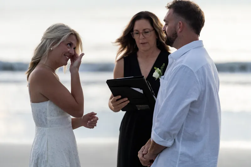 Jarah in her white dress wipes away tears as Brandon and their officiant look on during their beachside ceremony.
