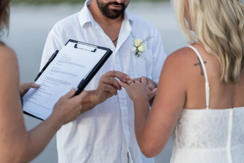 Brandon slides a ring onto Jarah's finger as their officiant holds the marriage document, with the ocean visible behind them.
