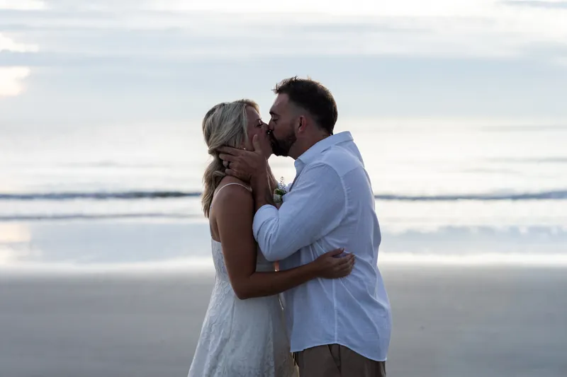 Jarah and Brandon kiss at the water's edge on Vilano Beach, waves rolling behind them under a cloudy sky.
