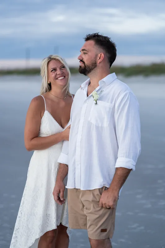Jarah laughs while looking at Brandon, who wears a white shirt and holds a small bouquet on Vilano Beach.