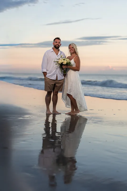 The couple stands on the reflective wet sand at sunset, Brandon holding Jarah's bouquet as the sky shifts to pink and gold.