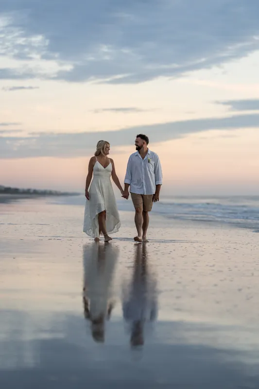 Walking barefoot through shallow water at Vilano Beach, they hold hands during the golden hour as the sun dips lower.