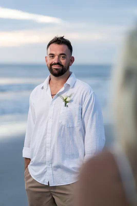 Brandon smiles at the camera wearing a white linen shirt with a boutonniere, ocean and sky visible behind him.