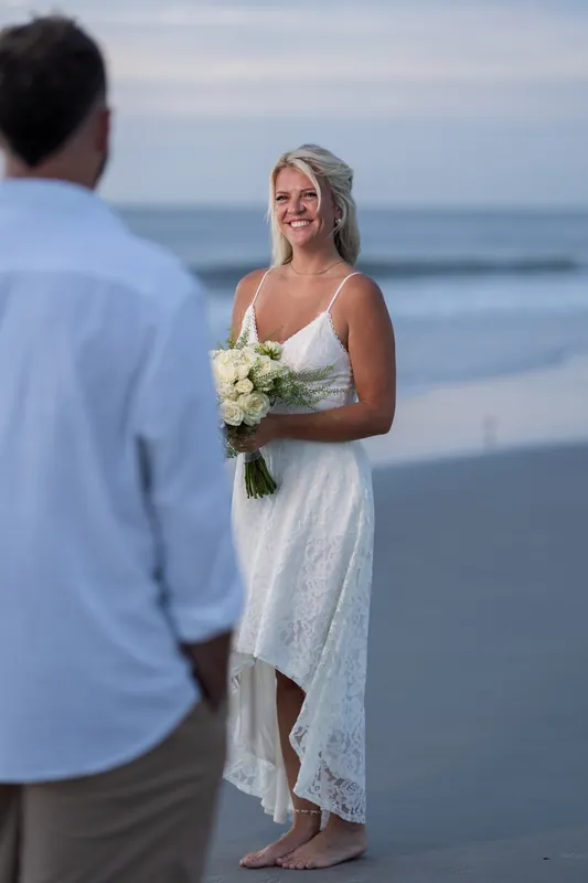 Jarah stands in a white lace dress holding a white bouquet while facing Brandon at the water's edge on Vilano Beach.