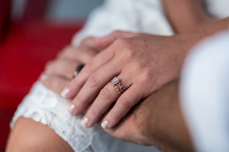 Brandon and Jarah's hands together display their wedding rings against red fabric and white lace.